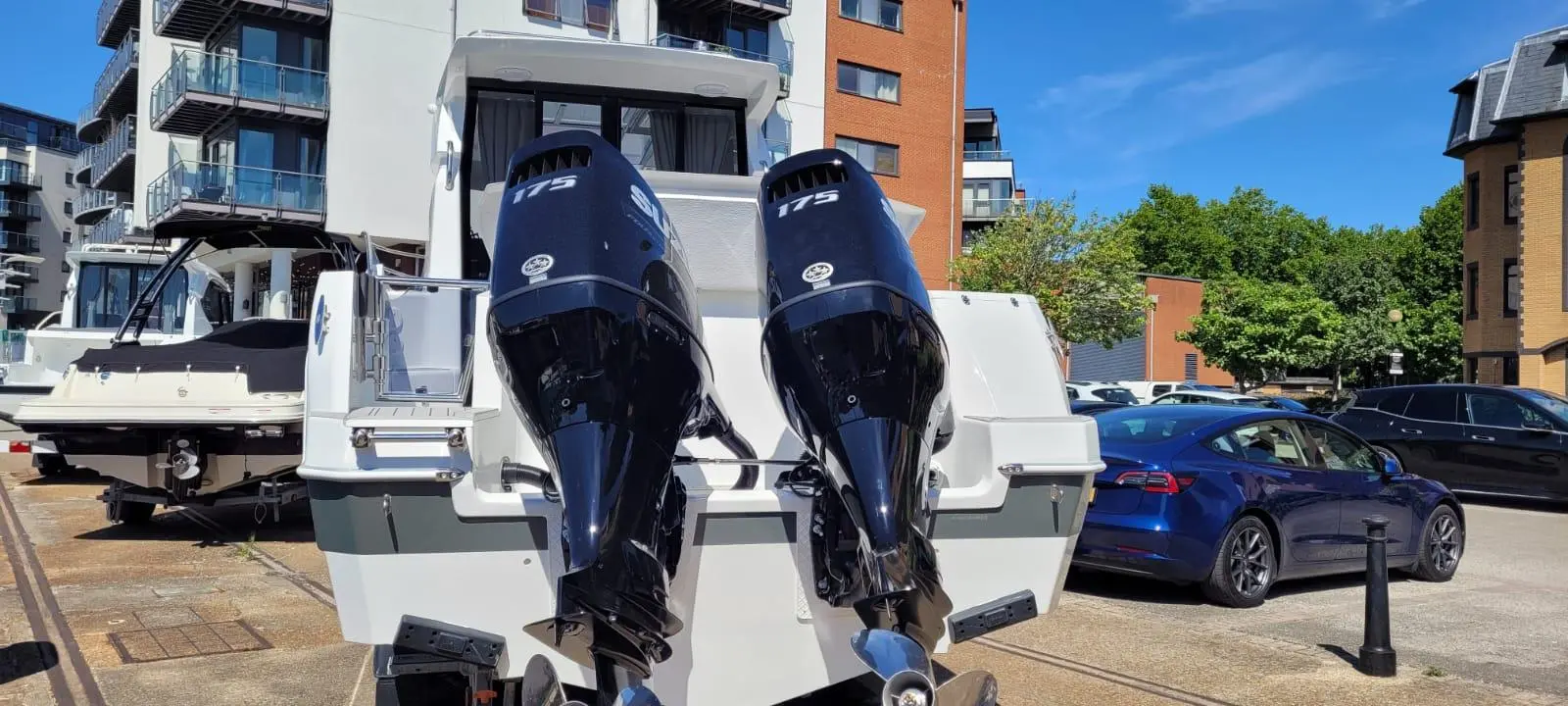 A white boat with two black outboard motors titled "175" docked near modern buildings and parked cars.